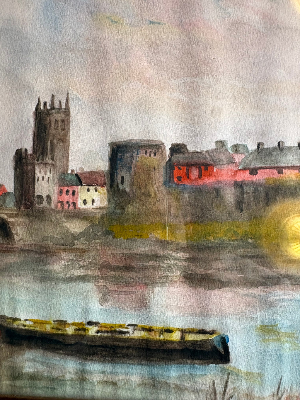 Riverside town detail showing a long boat, stone towers and red and pink buildings reflected in water.
