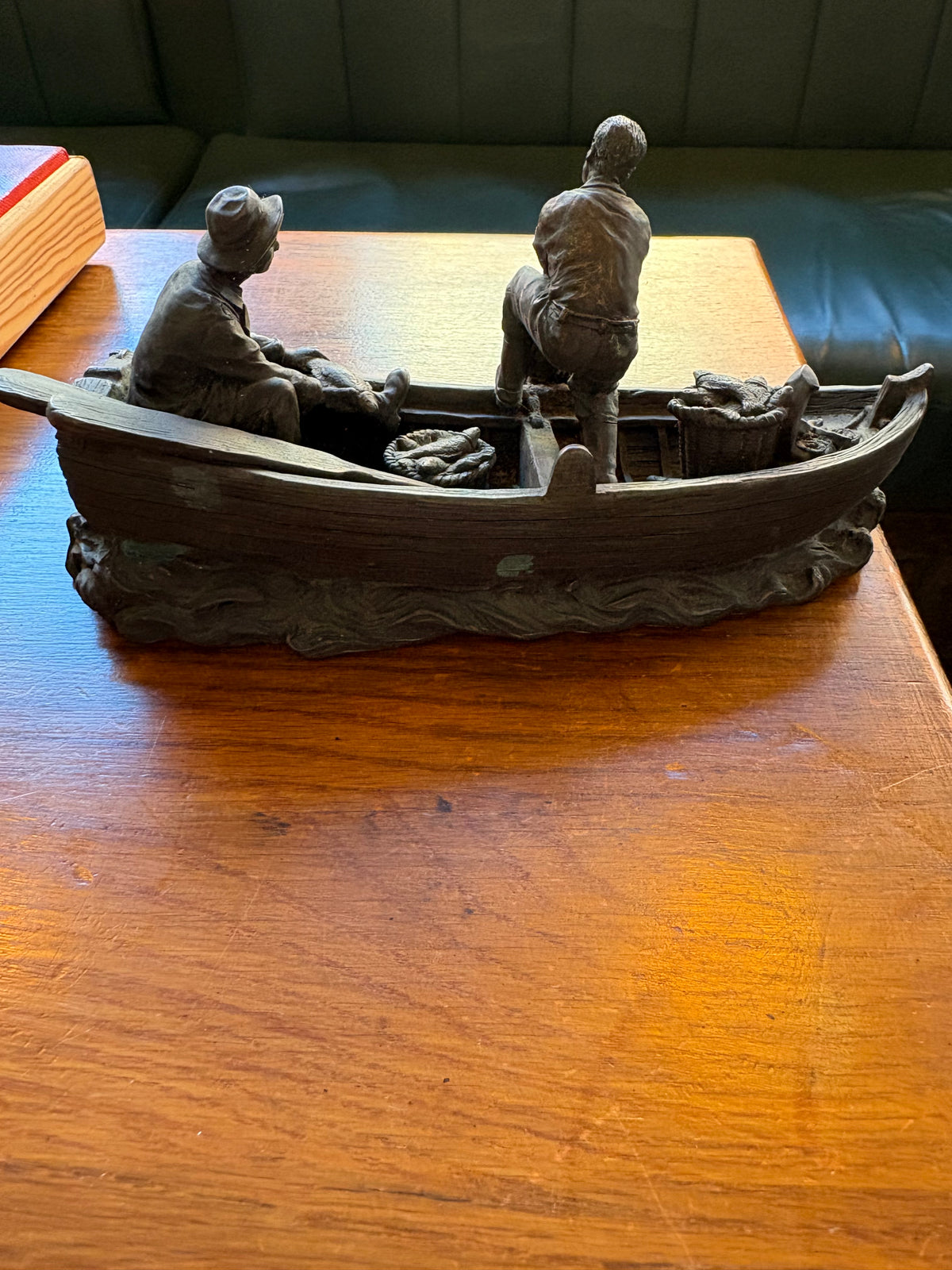 Rear side view of a bronze-coloured fishing boat sculpture with two fishermen, oars and baskets of fish