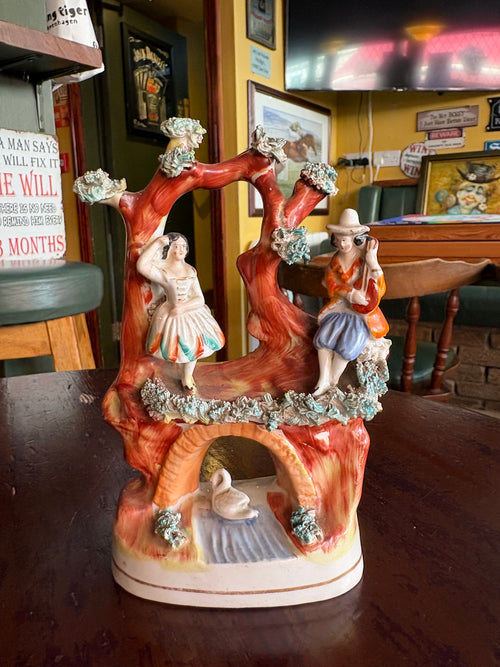 Colourful figurine group with a couple beside red tree trunks, textured foliage and a white swan beneath an arch