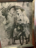 Framed monochrome picture of two children with flowers and a teddy bear beside a tricycle.